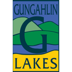 Gungahlin Lakes Golf Club logo: A tall rectangular badge with GUNGAHLIN in dark green serif capitals across a periwinkle blue sky, a large dark green serif letter G over light and dark green hills and a blue lake, and LAKES in dark green serif capitals on a yellow band beneath.